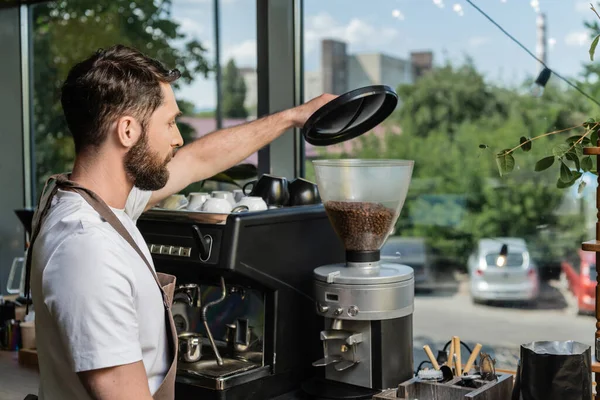 Vista lateral do barista no avental usando moedor de café enquanto trabalhava em café no fundo — Fotografia de Stock