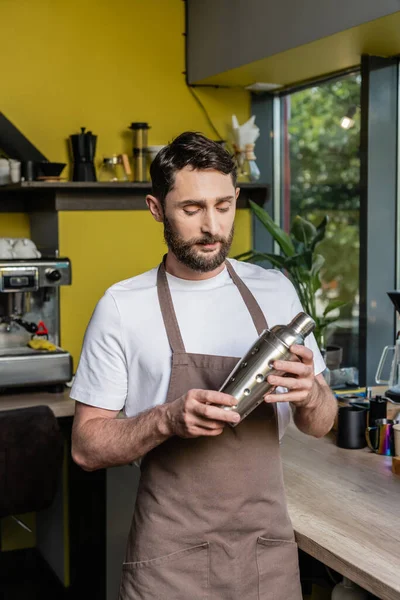 Bearded barista in apron holding shaker while standing near coffee machine in coffee shop — Stock Photo