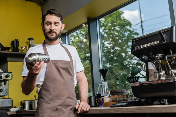 Barbudo barista en delantal usando agitador mientras trabaja cerca de la máquina de café en la cafetería - foto de stock