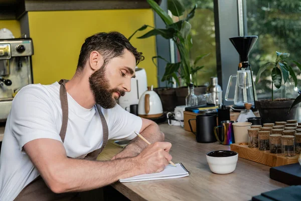 Barista barbudo em avental escrevendo no notebook perto de xícara e grãos de café no café — Fotografia de Stock