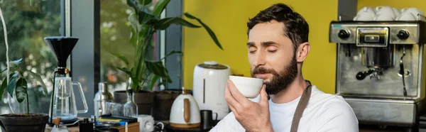 Bearded barista in apron smelling coffee in cup while working in coffee shop, banner — Stock Photo