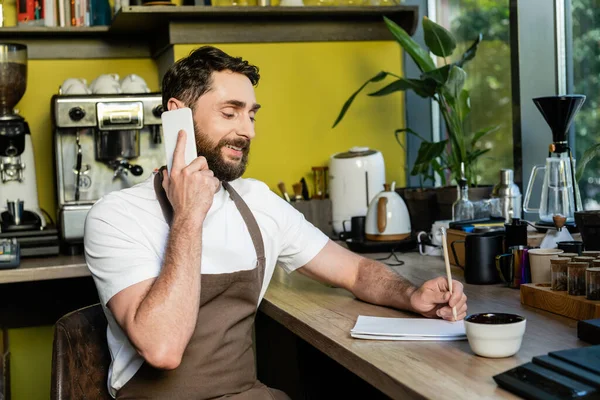 Barista sonriente en delantal hablando en el teléfono inteligente y escribiendo en el portátil en la cafetería - foto de stock