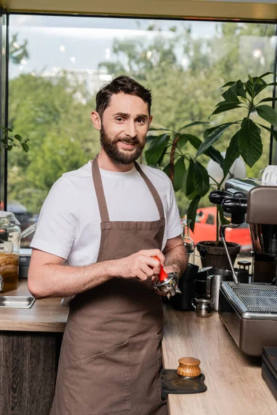 Lächelnder Barista blickt in die Kamera, während er im Coffeeshop Presse- und Kaffeemaschinenhalter hält — Stockfoto