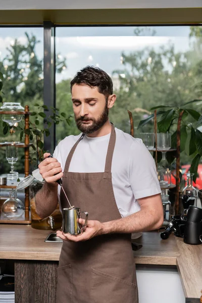 Bearded barista in apron holding milk frother and pitcher while working in coffee shop — Stock Photo
