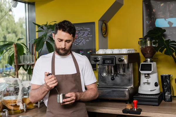 Barista in apron using milk frother and pitcher while working in coffee shop on background — Stock Photo