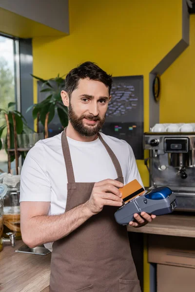 Smiling barista using payment terminal and credit card while looking at camera in coffee shop — Stock Photo