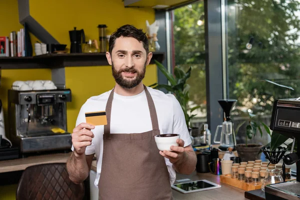 Lächelnder Barista in Schürze, der in die Kamera blickt, während er Kreditkarte und Tasse im Café hält — Stockfoto