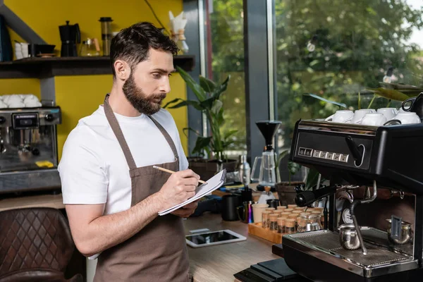 Бариста в фартуке, пишущая на блокноте, пользуясь кофемашиной и работая в кафе — стоковое фото