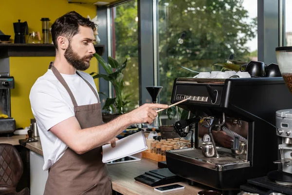 Barista holding notebook near coffee machine, smartphone and digital tablet in coffee shop — Stock Photo