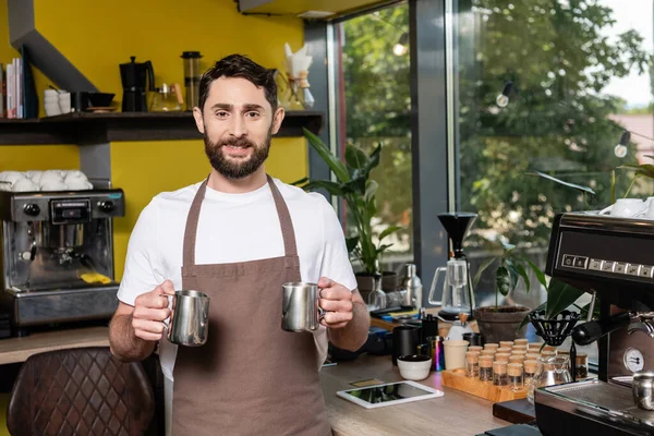 Cheerful barista holding pitchers while standing near coffee machine and digital tablet in cafe — Stock Photo
