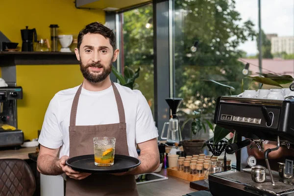Alegre barista en delantal sosteniendo limonada fría en vidrio y mirando a la cámara en la cafetería - foto de stock