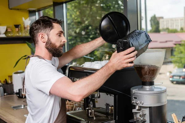 Vista lateral do barista no avental derramando grãos de café no moedor enquanto trabalhava no café — Fotografia de Stock