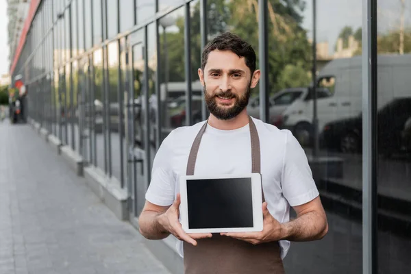 Усміхнений бариста в фартусі тримає цифровий планшет з порожнім екраном, стоячи на міській вулиці — стокове фото