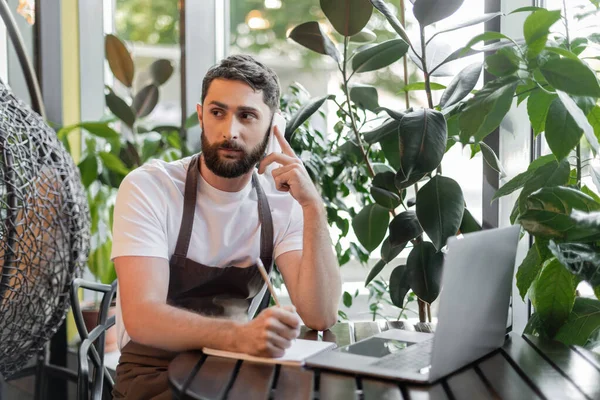 Barista parlando su smartphone e scrivendo su notebook vicino a computer portatile e piante in caffetteria — Foto stock