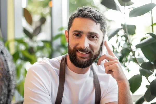 Portrait of positive bearded barista talking on smartphone and looking at camera in coffee shop — Stock Photo