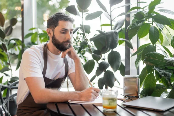 Barista in apron writing on notebook near lemonade and laptop on table in coffee shop — Stock Photo