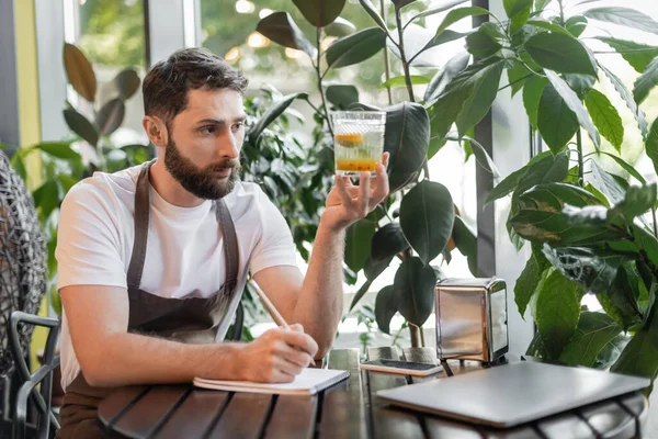 Barista in apron holding lemonade and writing on notebook near laptop on table in coffee shop — Stock Photo