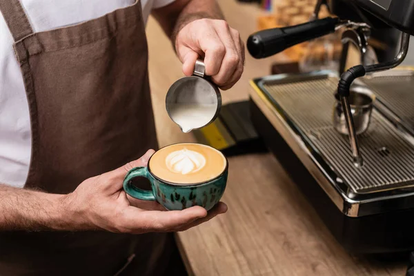 Cropped view of barista in apron pouring milk and making cappuccino near coffee machine in cafe — Stock Photo