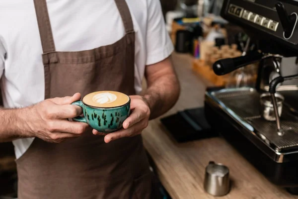 Vista recortada de barista en delantal sosteniendo la taza de capuchino mientras trabajaba en la cafetería - foto de stock