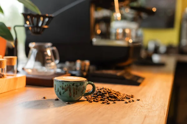Xícara de cappuccino perto de grãos de café na bancada perto de máquina de café borrada no café — Fotografia de Stock