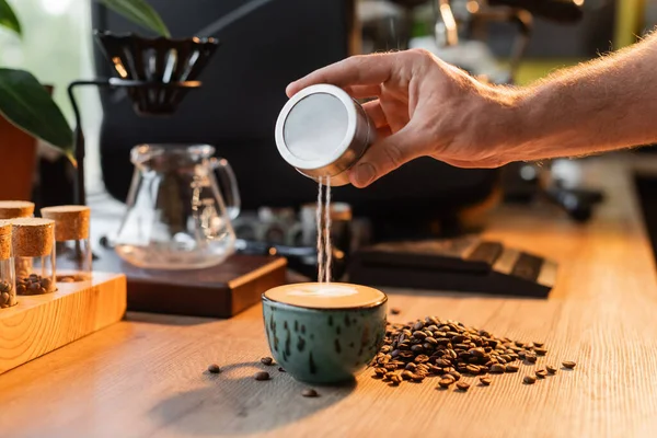 Vista cortada de barista derramando açúcar na xícara de cappuccino perto de grãos de café em luz no café — Fotografia de Stock