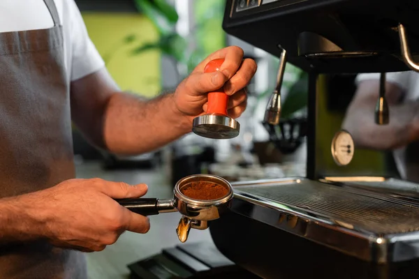 Vista cortada de barista no avental prendendo imprensa e café no suporte perto da máquina de café no café — Fotografia de Stock