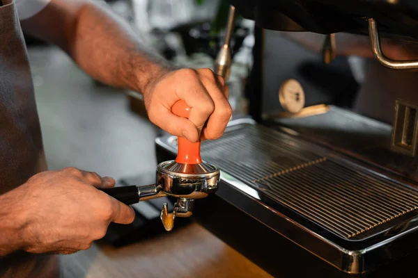Vista cortada de barista em suporte de imprensa avental segurando perto de máquina de café borrada no café — Fotografia de Stock