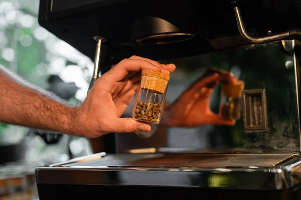 Vista cortada de barista segurando grãos de café em jarra perto de máquina de café borrada no café — Fotografia de Stock