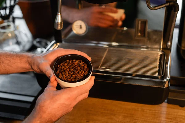Cropped view of barista holding cup with coffee beans near blurred coffee machine in cafe — Stock Photo