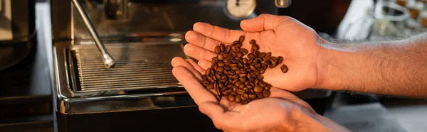 Vista recortada de barista sosteniendo granos de café cerca de la máquina de café borrosa en la cafetería, pancarta - foto de stock