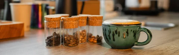 Cup of cappuccino near coffee beans in jars on wooden worktop in blurred coffee shop, banner — Stock Photo