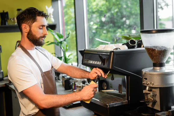 Barbudo barista limpieza de la máquina de café boquilla con trapo mientras trabaja en la cafetería - foto de stock