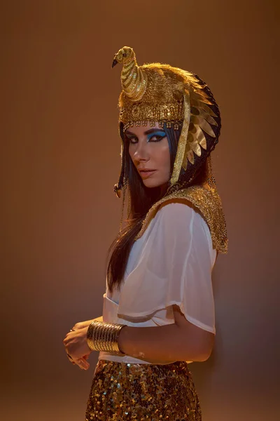 Mujer con estilo en traje egipcio y tocado mirando a la cámara mientras posando aislado en marrón - foto de stock