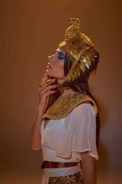 Frau in ägyptischer Kleidung und Kopfbedeckung berührt Kinn, während sie auf braunem Hintergrund posiert — Stockfoto