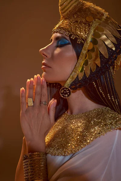 Вид сбоку женщины в египетской одежде и головной убор делает молитвенные руки жестом на коричневом фоне — стоковое фото