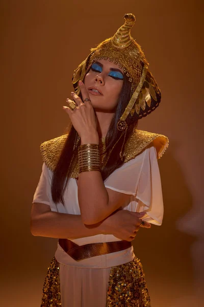 Mulher elegante com maquiagem ousada e trajes egípcios posando com olhos fechados no fundo marrom — Fotografia de Stock