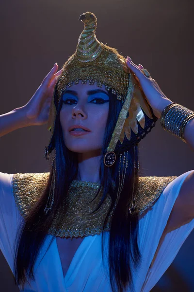 Mujer atractiva tocando el tocado egipcio y mirando a la cámara en luz azul sobre fondo marrón - foto de stock