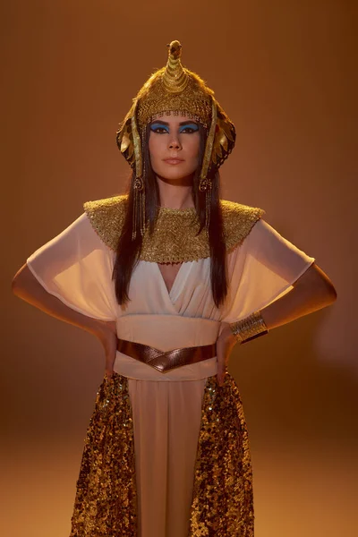 Mulher morena elegante na cobertura para a cabeça egípcia e olhar de mãos dadas em quadris no fundo marrom — Fotografia de Stock