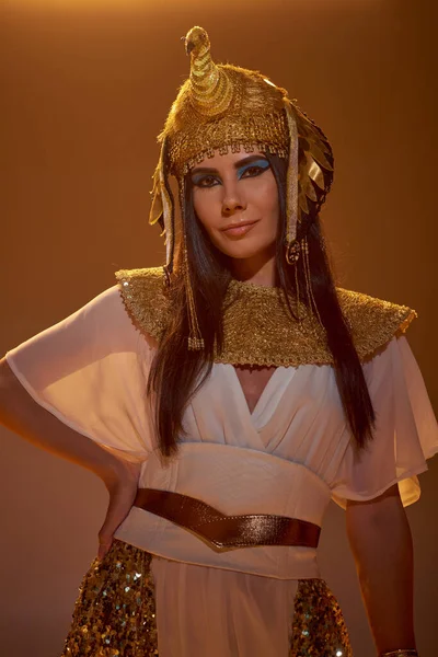 Брюнетка в древнеегипетском костюме и головном уборе, смотрящая на камеру, изолированную от коричневого — стоковое фото