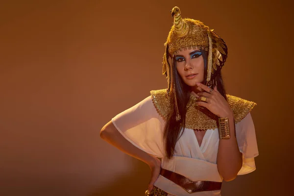 Стильная женщина в египетской одежде и головной убор трогательный подбородок и позирование на коричневом фоне — стоковое фото