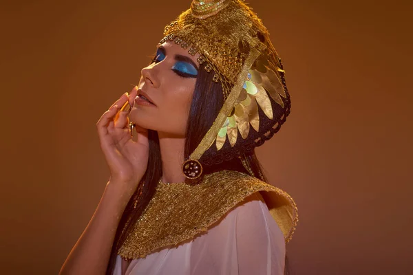 Portrait of woman with bold makeup and egyptian headdress touching cheek isolated on brown — Stock Photo