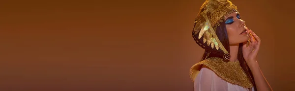 Брюнетка жінка з сміливим макіяжем, одягнена в єгипетський головний убір, позує ізольовано на коричневому, банер — Stock Photo