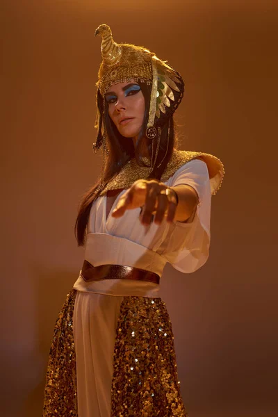Mulher de traje egípcio apontando com o dedo para a câmera e em pé sobre fundo marrom com luz — Fotografia de Stock