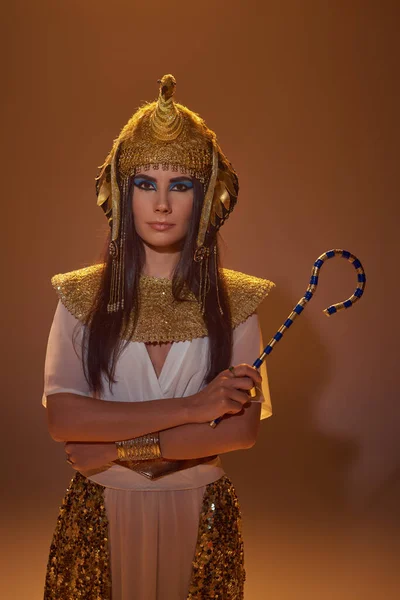Brunette woman in egyptian attire and headdress holding crook and standing on brown background — Stock Photo