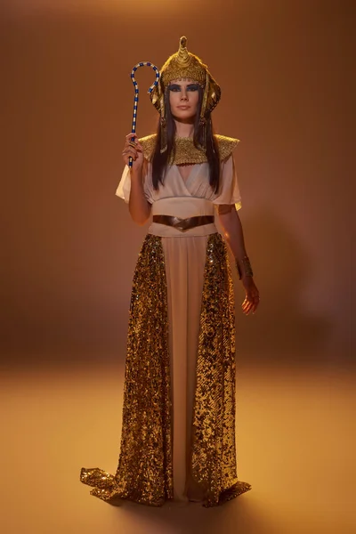 Full length of stylish woman in egyptian attire holding crook and standing on brown background — Stock Photo