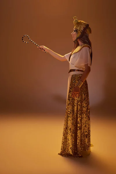 Вид збоку елегантної жінки в єгипетському вбранні тримає криву, стоячи на коричневому фоні — стокове фото