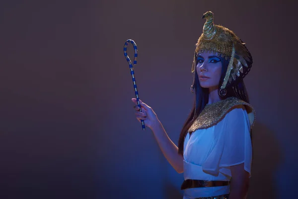 Brunette woman in egyptian attire and headdress holding crook on brown background with blue light — Stock Photo