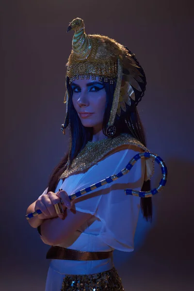 Portrait of woman in egyptian look holding crook and posing on brown background with blue light — Stock Photo
