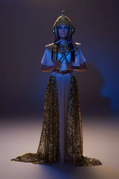 Pleine longueur de femme élégante dans le regard égyptien tenant escroc et fléau sur brun avec lumière bleue — Photo de stock