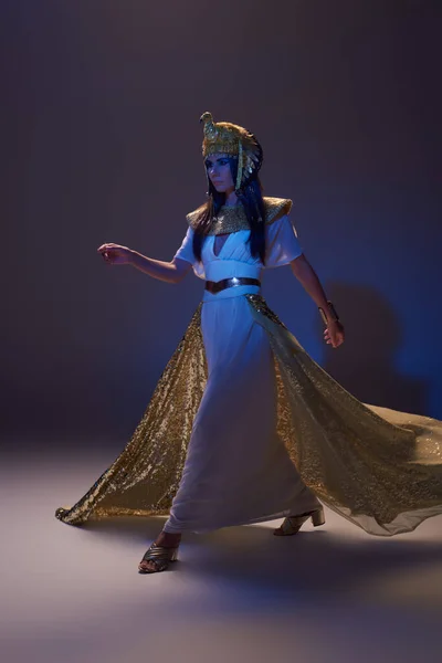 Piena lunghezza della donna in look egiziano e copricapo a piedi su sfondo marrone con luce blu — Foto stock